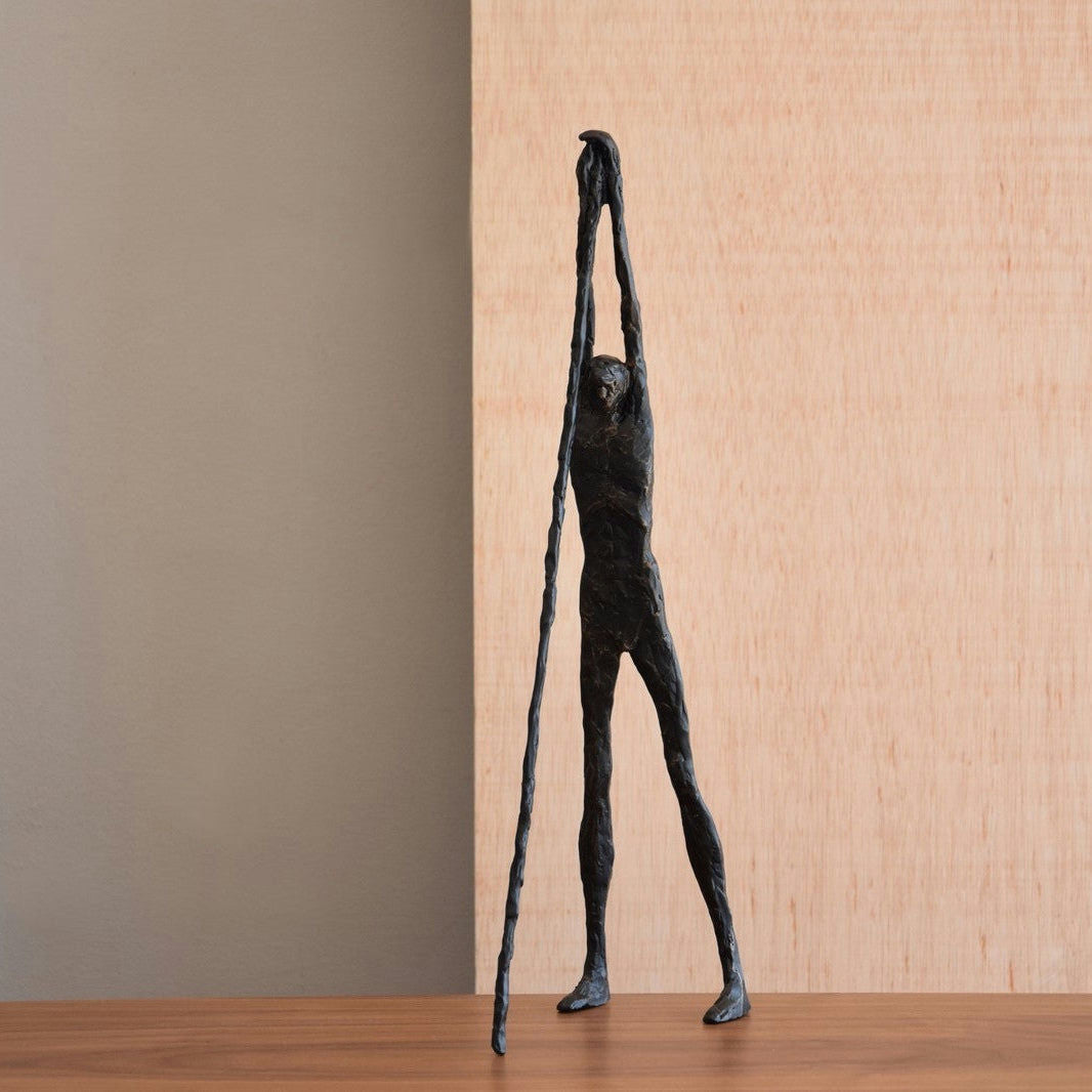 La figura decorativa de metal en bronce envejecido N2 presenta una figura alta y delgada con brazos alargados que se extienden hacia arriba, de pie con gracia sobre una superficie de madera, ideal para los entusiastas del diseño de interiores que buscan elegancia y arte.