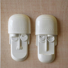  Dos Máscaras Decorativas en Cerámica Blanco Brillante descansan sobre un fondo de madera. Cada una presenta un rostro estilizado con partes superiores que parecen cascos, narices redondas y ojos cerrados, lo que agrega un encanto enigmático a cualquier colección artística.