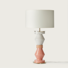  Lámpara de mesa moderna con pantalla blanca y base escalonada que alterna segmentos rosas y blancos sobre un fondo blanco.