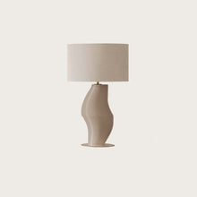  Una moderna Lámpara de mesa Luet (Cristal o Metal) con una base texturizada y entallada y una pantalla circular lisa de color blanquecino sobre un fondo neutro, que muestra una iluminación elegante.