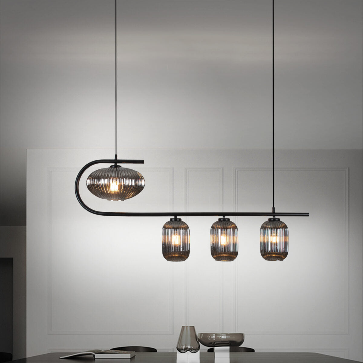 La lámpara de techo en acero diseño industrial B-Kairo 4, que consta de cuatro luces colgantes con pantallas de alambre en una luminaria curva, cuelga sobre una mesa oscura, brindando iluminación difusa junto a un pequeño vaso y otro objeto.