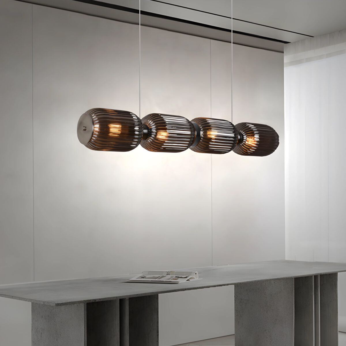La Lámpara de techo en acero y vidrio Kairo 4 presenta cuatro pantallas de vidrio acanalado que cuelgan sobre una mesa gris en una habitación minimalista, ofreciendo una iluminación uniforme.