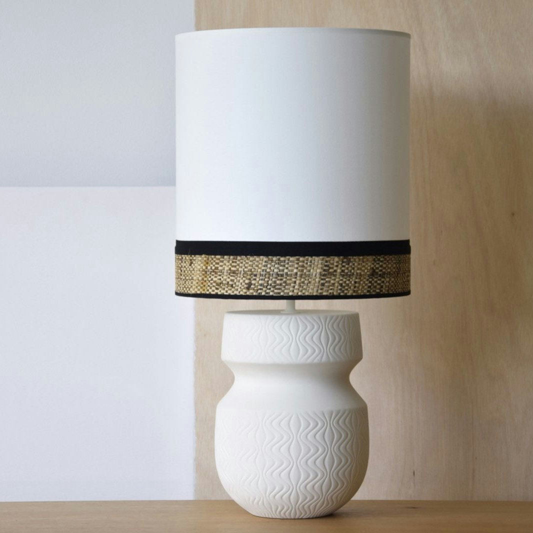 La Lámpara de Mesa Cerámica - Eva, que cuenta con una base de cerámica texturizada blanca y una gran Pantalla Eva cilíndrica, reposa sobre una superficie de madera. La pantalla tiene una banda decorativa cerca de su borde inferior.