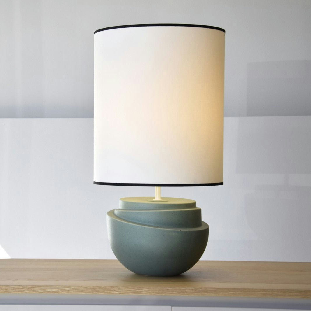 La Lámpara de Mesa Cerámica - Julieta presenta una moderna pantalla cilíndrica pálida y una base geométrica apilada en verde apagado, sobre una superficie de madera clara, ideal para realzar cualquier diseño de interiores.