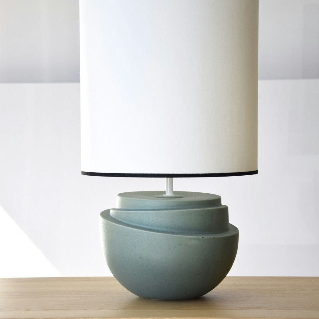 La Lámpara de Mesa Cerámica - Julieta presenta una base verde azulado mate y una pantalla cilíndrica alta, elegantemente situada sobre una superficie de madera.