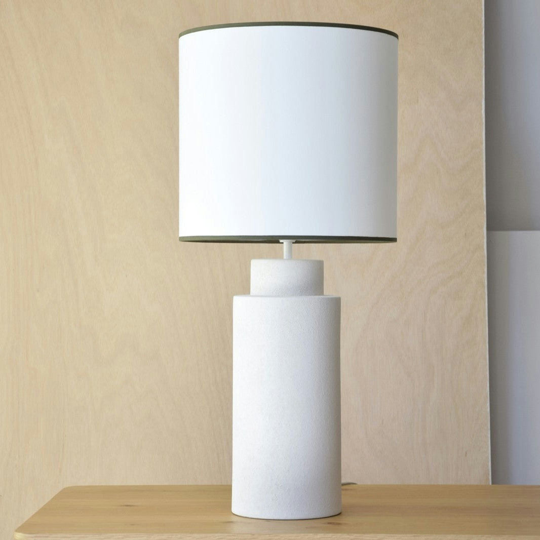 La Lámpara de Mesa Cerámica MID, una moderna lámpara de mesa blanca con base cilíndrica y pantalla redonda grande en diseño Mid Century, reposa sobre una superficie de madera sobre un fondo de madera sencillo.