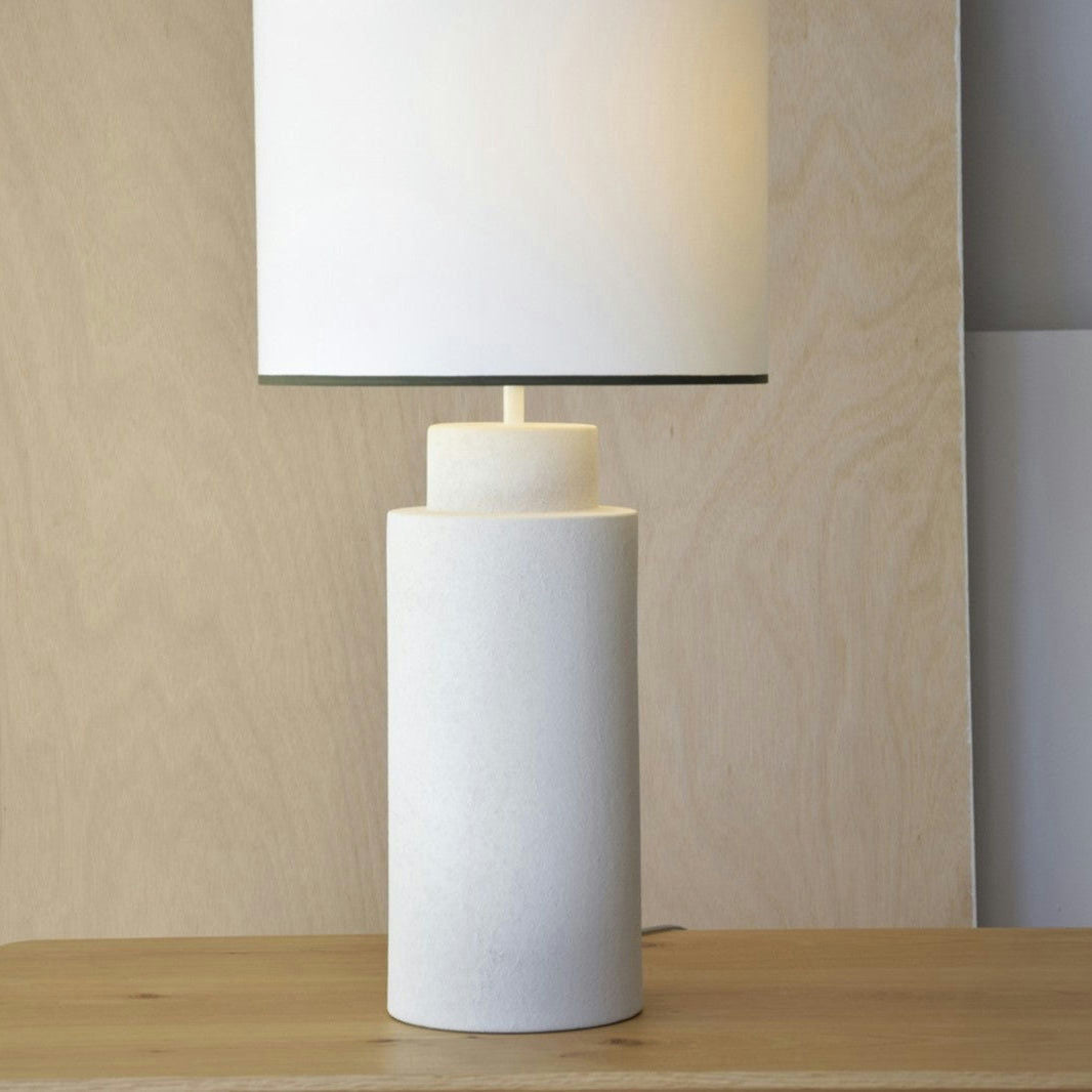 La Lámpara de Mesa Cerámica - MID es una lámpara de mesa moderna con una base cilíndrica y una pantalla rectangular. Presenta una pantalla de algodón blanco y reposa elegantemente sobre madera, contra un fondo claro, adoptando el encanto del diseño de mediados de siglo.