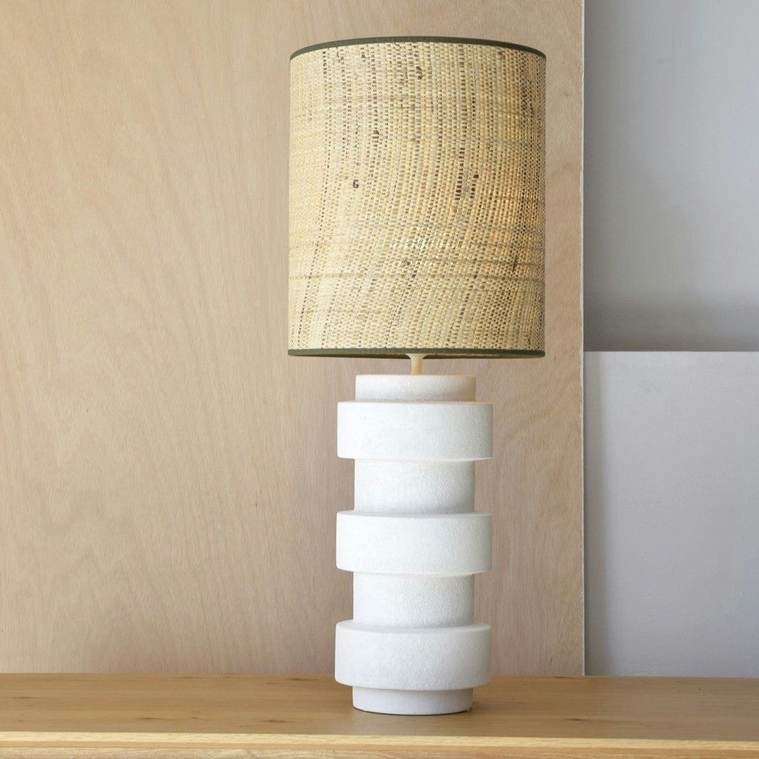 Una Lámpara de Mesa Cerámica - Vega con una base cilíndrica blanca texturizada y una pantalla de rafia natural se asienta sobre una superficie de madera, exudando un encanto de estilo rústico.