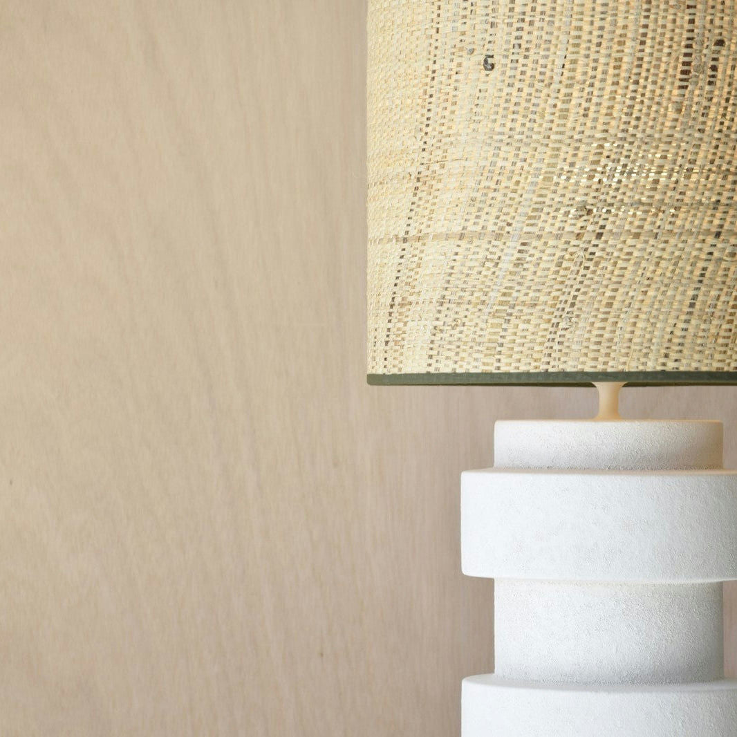 La Lámpara de Mesa Cerámica - Vega, con su pantalla Pantalla tejida en color beige sobre una base texturizada en blanco, complementa una pared en color beige a juego, irradiando el encanto del Estilo Rústico.