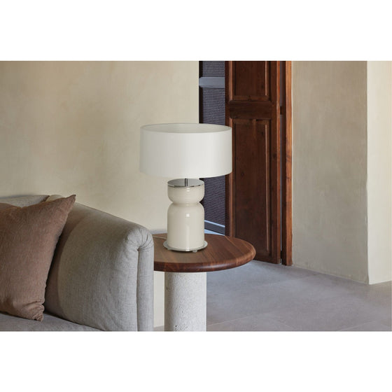 Una Lámpara de Mesa moderna se colocó sobre una mesa auxiliar redonda de madera junto a un sofá gris en una habitación minimalista con una pared beige y una puerta de madera, con iluminación versátil.