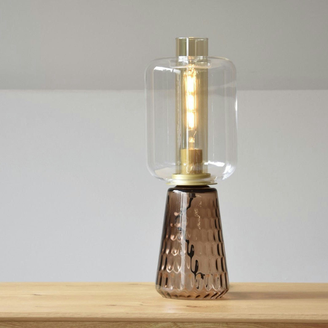 La Lámpara de Mesa Led Colección Candil Vidrio Ámbar de Lucerna, con una pantalla de vidrio ámbar transparente y una base de bronce texturizada, se asienta elegantemente sobre una superficie de madera con un elegante diseño moderno.