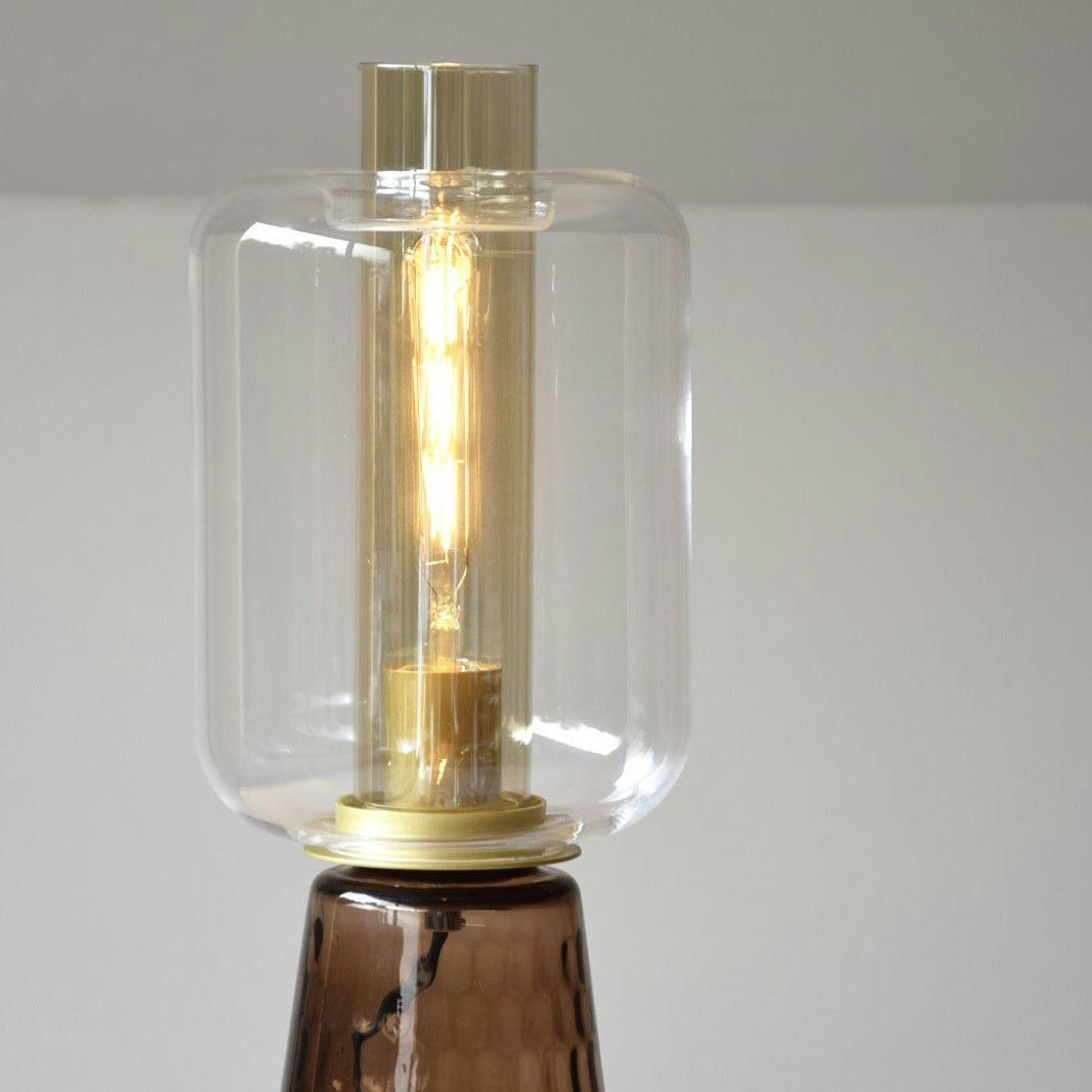 Una Lámpara de Mesa Led Colección Candil Vidrio Ámbar - Lucerna encendida con una pantalla de vidrio transparente, una bombilla cilíndrica y un detalle de base metálica se coloca sobre un fondo liso.
