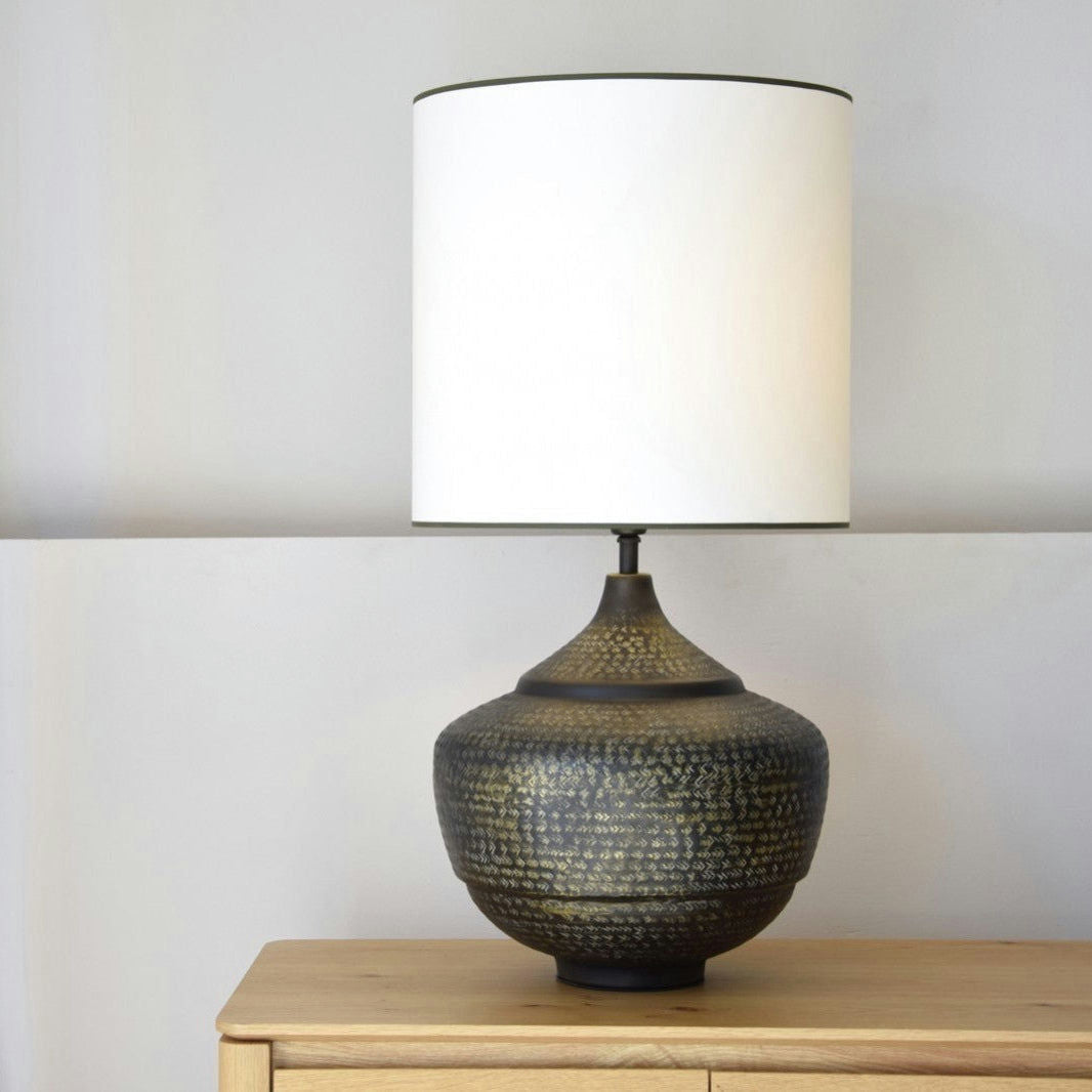 La Lámpara de Mesa Metal N.1, con una base de cerámica oscura texturizada y una pantalla cilíndrica blanca, se asienta sobre una superficie de madera, complementando perfectamente cualquier diseño de interiores.