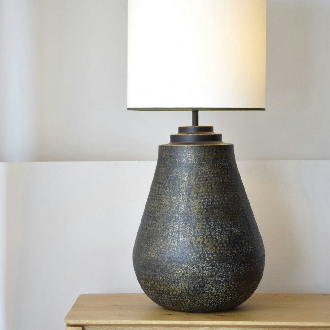 La Lámpara de Mesa Metal N.10, con una base oscura y texturizada y una pantalla cilíndrica blanca, se asienta elegantemente sobre una superficie de madera clara contra una pared neutra, mostrando una exquisita iluminación artesanal.