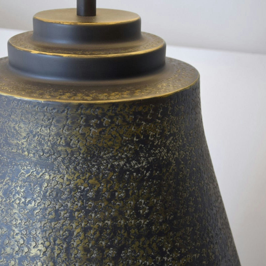 Primer plano de la pantalla de metal oscuro texturizado de la Lámpara de Mesa Metal N.10, con tonos dorados y negros. El elegante diseño se estrecha desde una parte superior cilíndrica hasta una parte inferior más ancha, mostrando la iluminación artesanal.