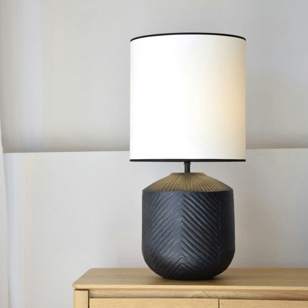 La Lámpara de Mesa Metal N.2 - Ferreus, con una pantalla cilíndrica blanca y una base oscura texturizada con artesanía en hierro, está elegantemente colocada sobre una superficie de madera contra una pared de color claro.