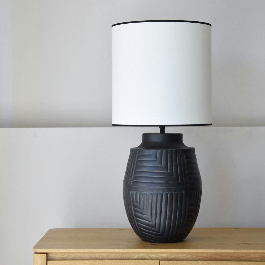 La Lámpara de Mesa Metal N7 - Luxor, con una base de cerámica texturizada negra y una pantalla cilíndrica blanca, descansa elegantemente sobre una superficie de madera contra una pared lisa, realzando el diseño de interiores.