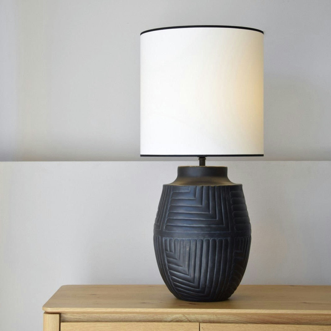 La Lámpara de Mesa Metal N7 - Luxor, una lámpara de cerámica negra con un patrón geométrico texturizado, se encuentra elegantemente sobre una mesa de madera. Su pantalla cilíndrica blanca contrasta con la pared gris claro, agregando un toque de diseño de interiores.
