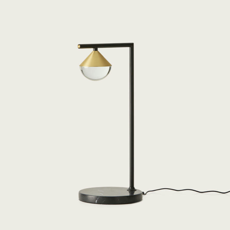 La Lámpara de Mesa Nino cuenta con un estilo moderno con un soporte de metal negro, una pantalla cónica dorada y una cubierta de vidrio redonda sobre una base circular negra. Su diseño contemporáneo agrega un toque elegante a cualquier espacio, y se completa con un cable de alimentación.