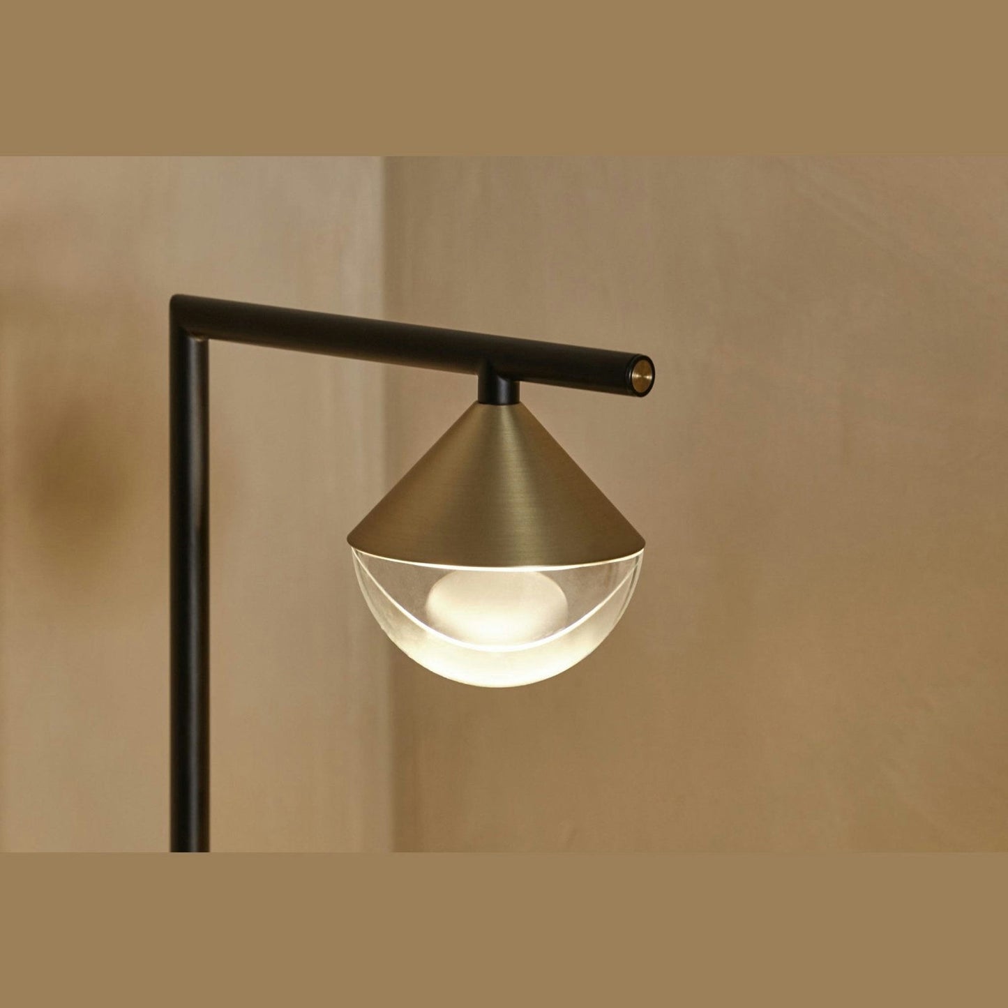 La Lámpara de Mesa Nino presenta una pantalla metálica en forma de cono y un difusor de vidrio sobre un brazo negro, presentando un diseño contemporáneo contra una pared beige.