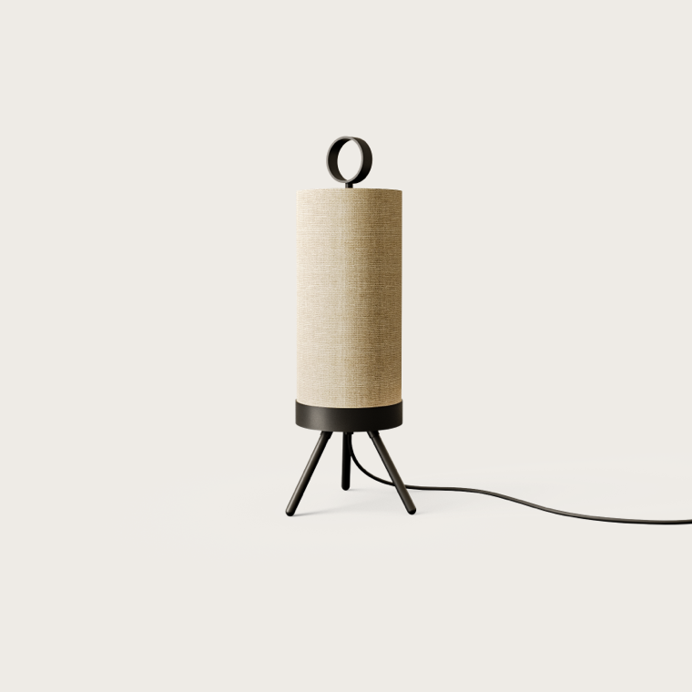 La Lámpara de Mesa Nooi es una pieza cilíndrica alta de color beige con una base negra y patas de trípode, completa con un asa en forma de bucle en la parte superior. Un cable de alimentación negro emerge de la base, lo que se suma a su diseño minimalista escandinavo sobre un fondo blanco liso.