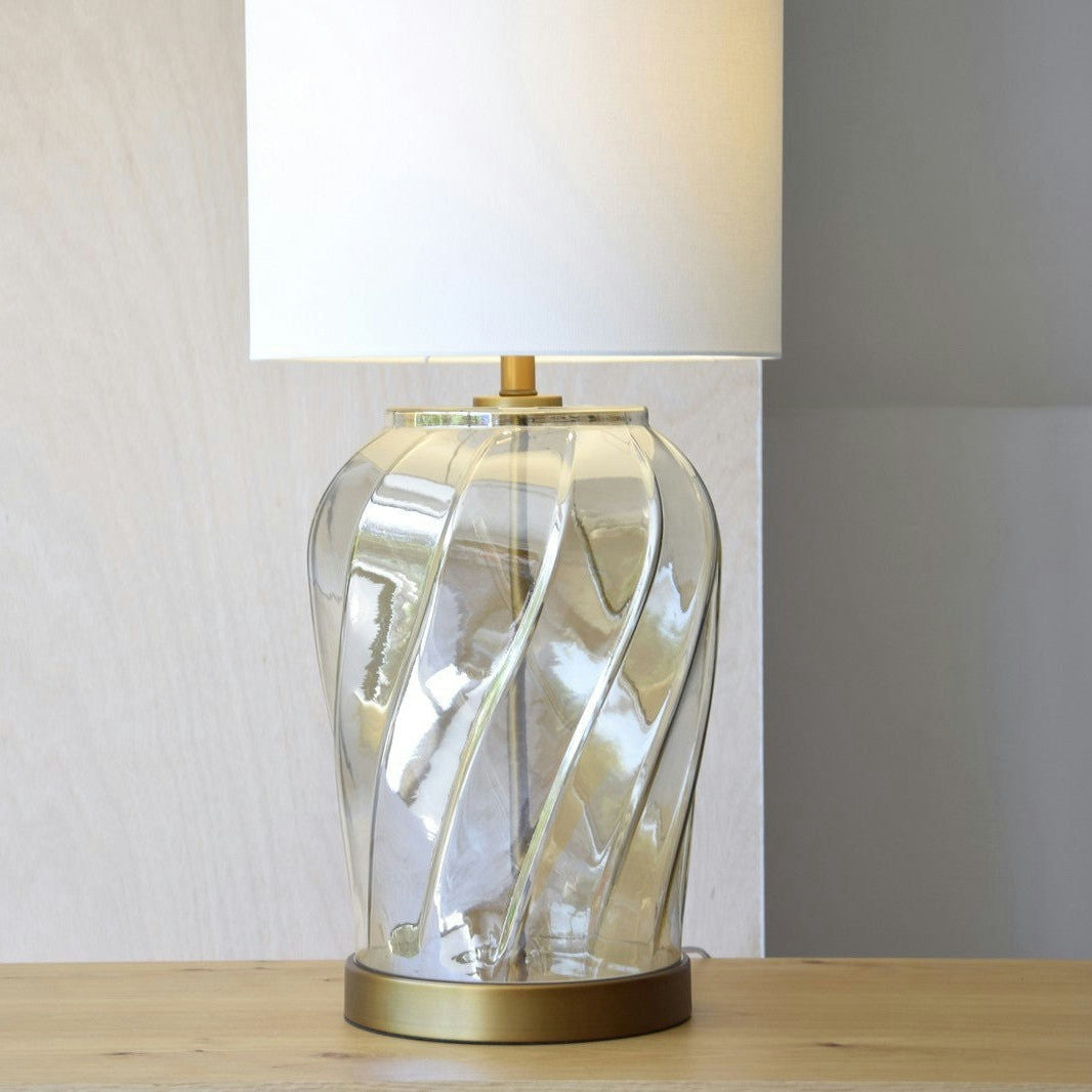 La Lámpara de Mesa Vidrio Ámbar Twist, colocada sobre una mesa de madera, presenta un diseño en espiral con una pantalla blanca y una base dorada. Esta lámpara contemporánea realza maravillosamente la decoración y el interiorismo.
