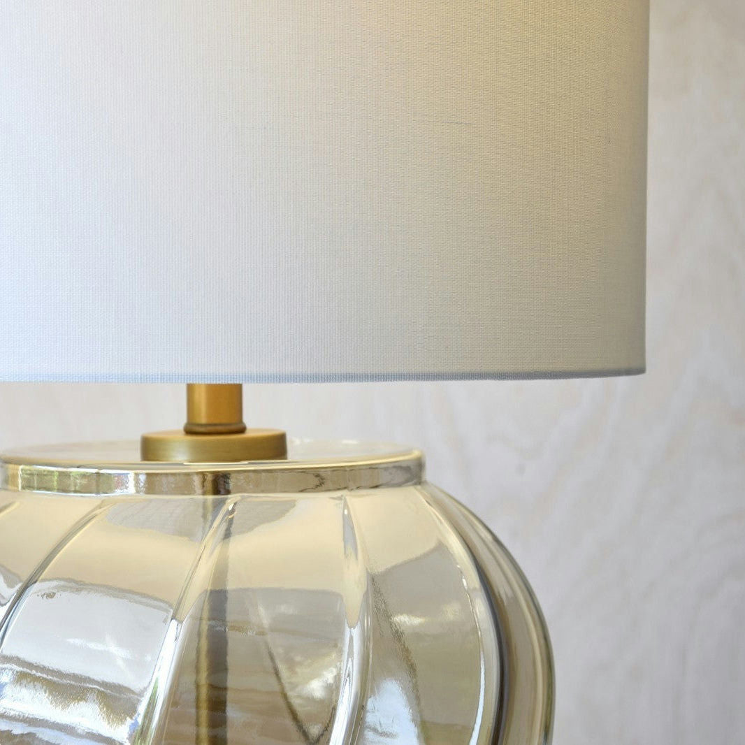 La Lámpara de Mesa Vidrio Ámbar Twist presenta una base de vidrio texturizado con una pantalla de tela beige, ideal para agregar estilo contemporáneo a cualquier habitación.