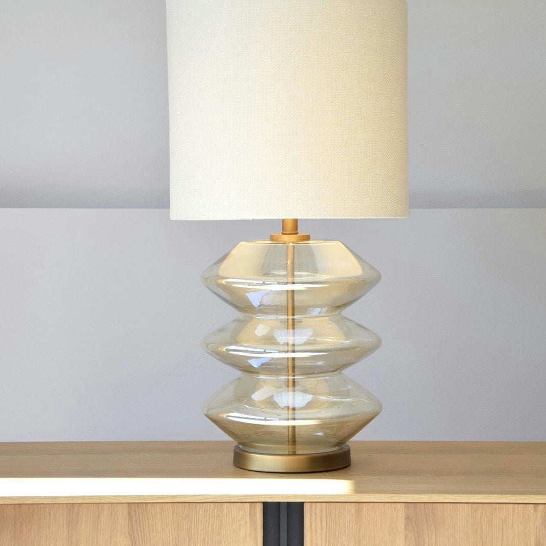 La Lámpara de Mesa Zig Zag de Vidrio Ámbar / Blanco, con su escultural base de vidrio ámbar transparente y pantalla beige, se asienta elegantemente sobre un gabinete de madera.