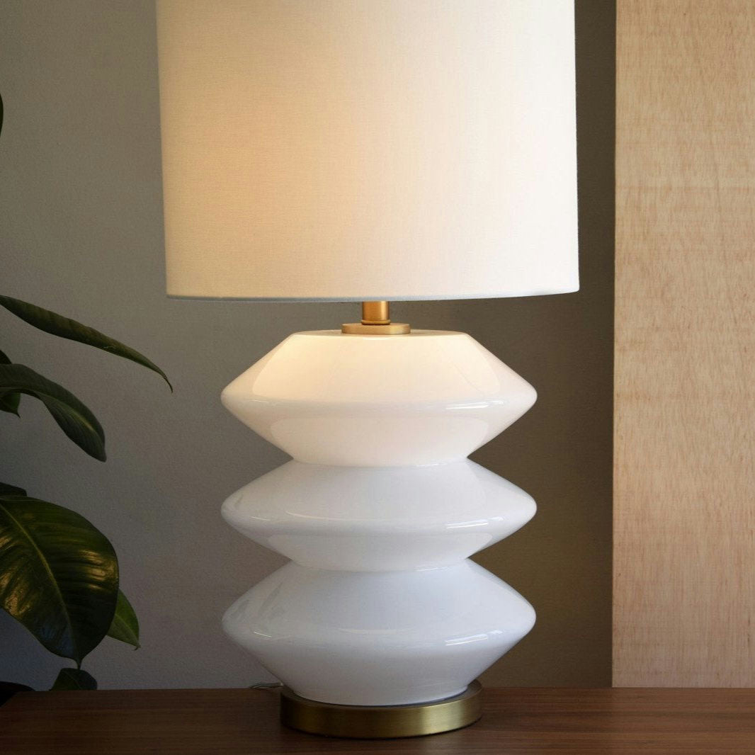La Lámpara de Mesa Zig Zag de Vidrio Ámbar/Blanco cuenta con una base de cerámica geométrica apilada con una pantalla de tela cilíndrica, que le agrega un toque de diseño contemporáneo. Descansa sobre una superficie de madera junto a plantas de hojas verdes para realzar la atmósfera acogedora.