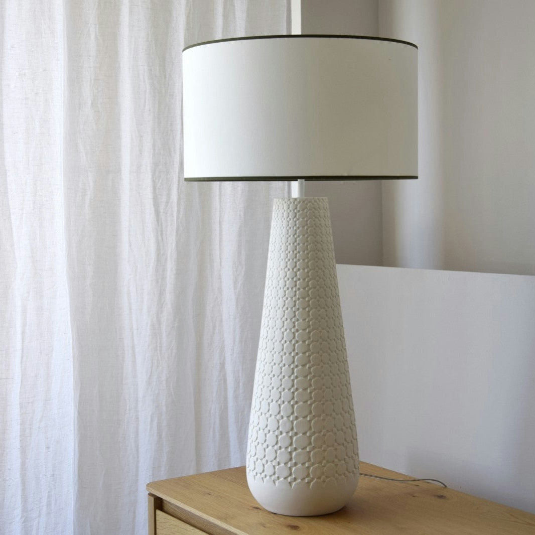 La Lámpara de Mesa de Cerámica Claudia con Pantalla tiene un diseño innovador blanco con base cónica texturizada de cerámica natural y pantalla redonda, ubicada sobre una superficie de madera junto a cortinas.