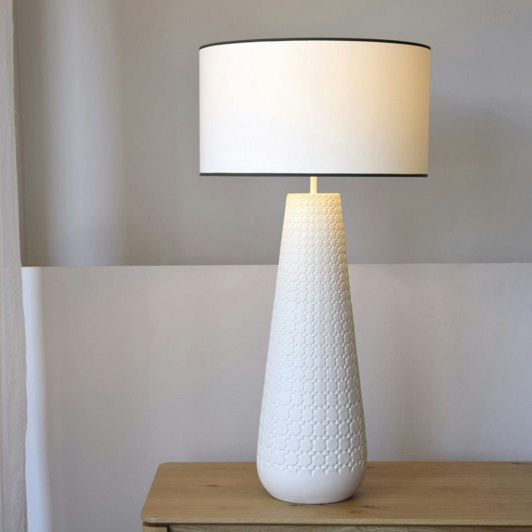 La Lámpara de Mesa de Cerámica Claudia con Pantalla presenta una base cónica texturizada hecha de cerámica de chacota natural y una pantalla redonda blanca colocada sobre una superficie de madera.