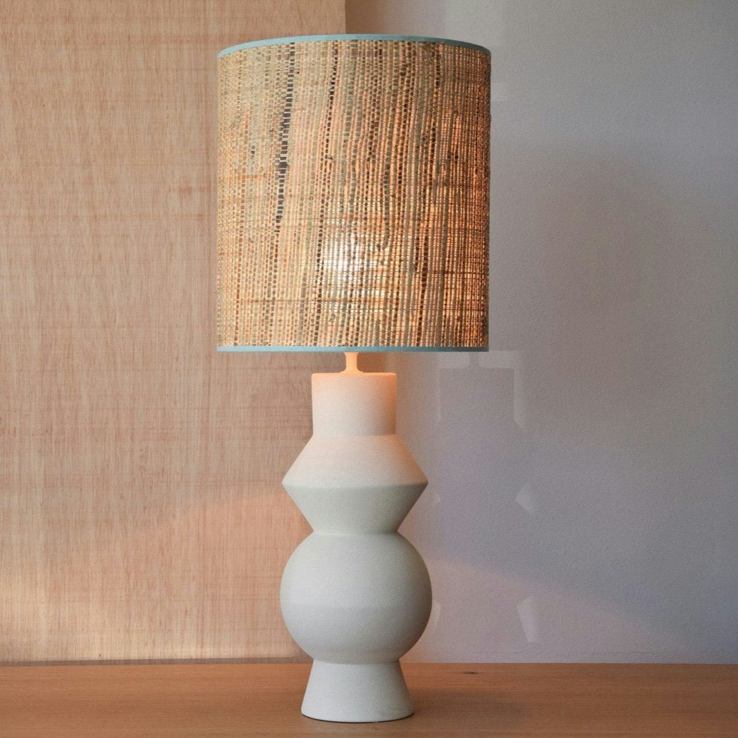La Lámpara de Mesa de Cerámica Forma Blanco Crudo cuenta con una pantalla cilíndrica beige texturizada hecha de pantalla rafia natural, colocada sobre una base geométrica blanca de cerámica blanco crudo, elegantemente colocada sobre una superficie de madera contra un fondo neutro.