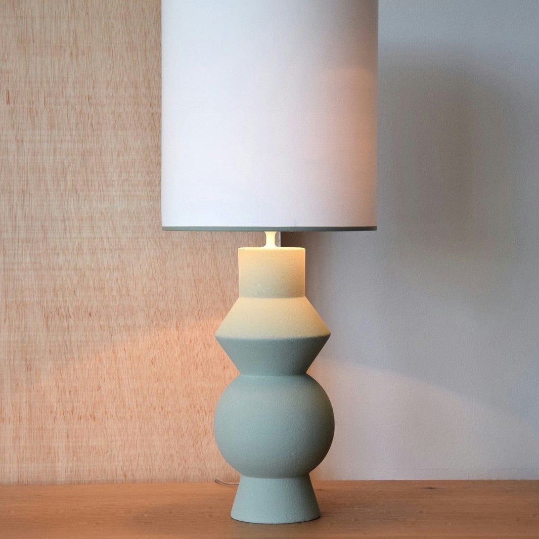 La Lámpara de Mesa de Cerámica Verde Forma, con una base geométrica y una sencilla pantalla blanca, se asienta sobre madera sobre un fondo neutro, mostrando su diseño contemporáneo.