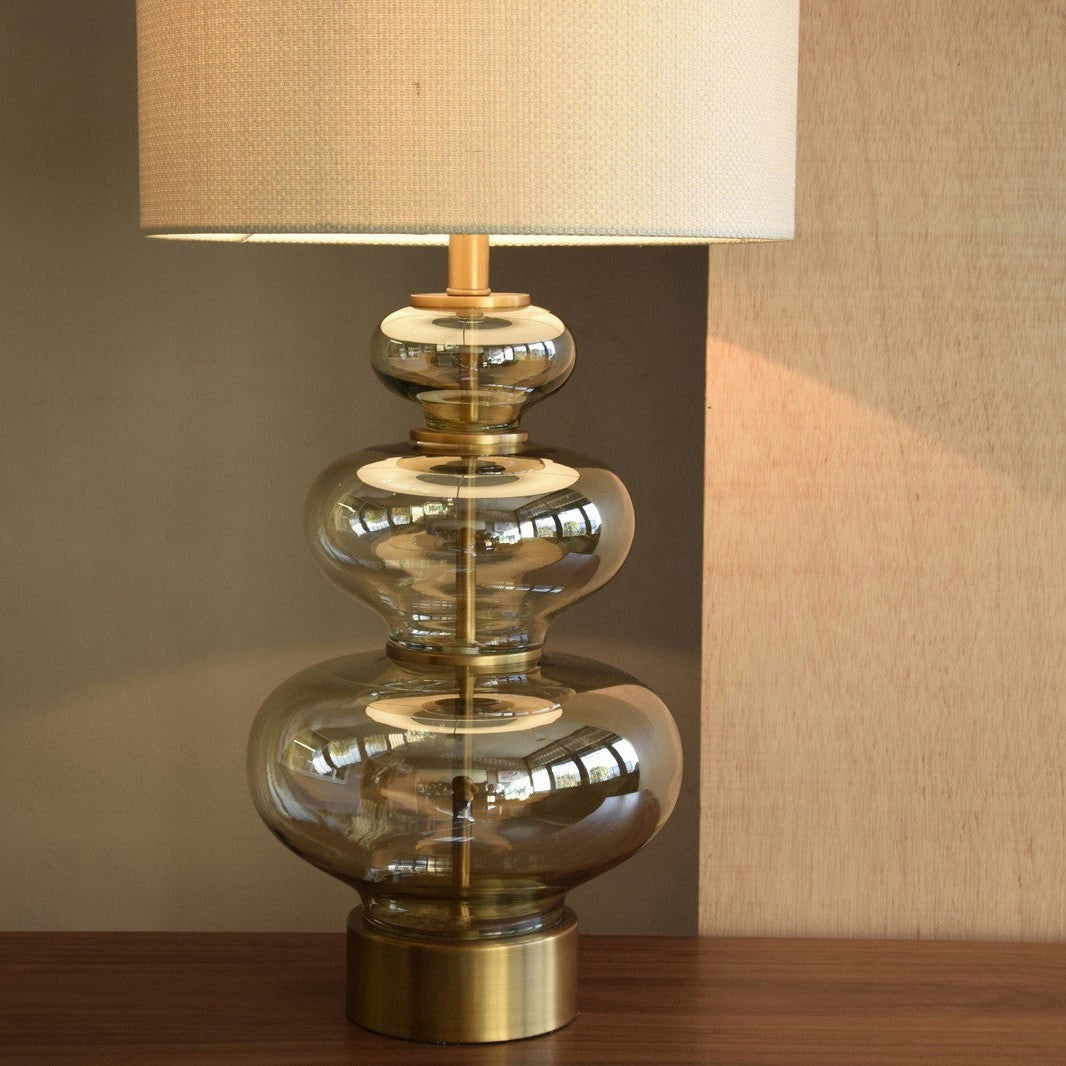 La Lámpara de Mesa de Vidrio Ámbar Calabaza, con una base de vidrio apilado en formas bulbosas y una pantalla de tela beige, se asienta elegantemente sobre una superficie de madera, presentando una elegante base dorada envejecida para mayor encanto.