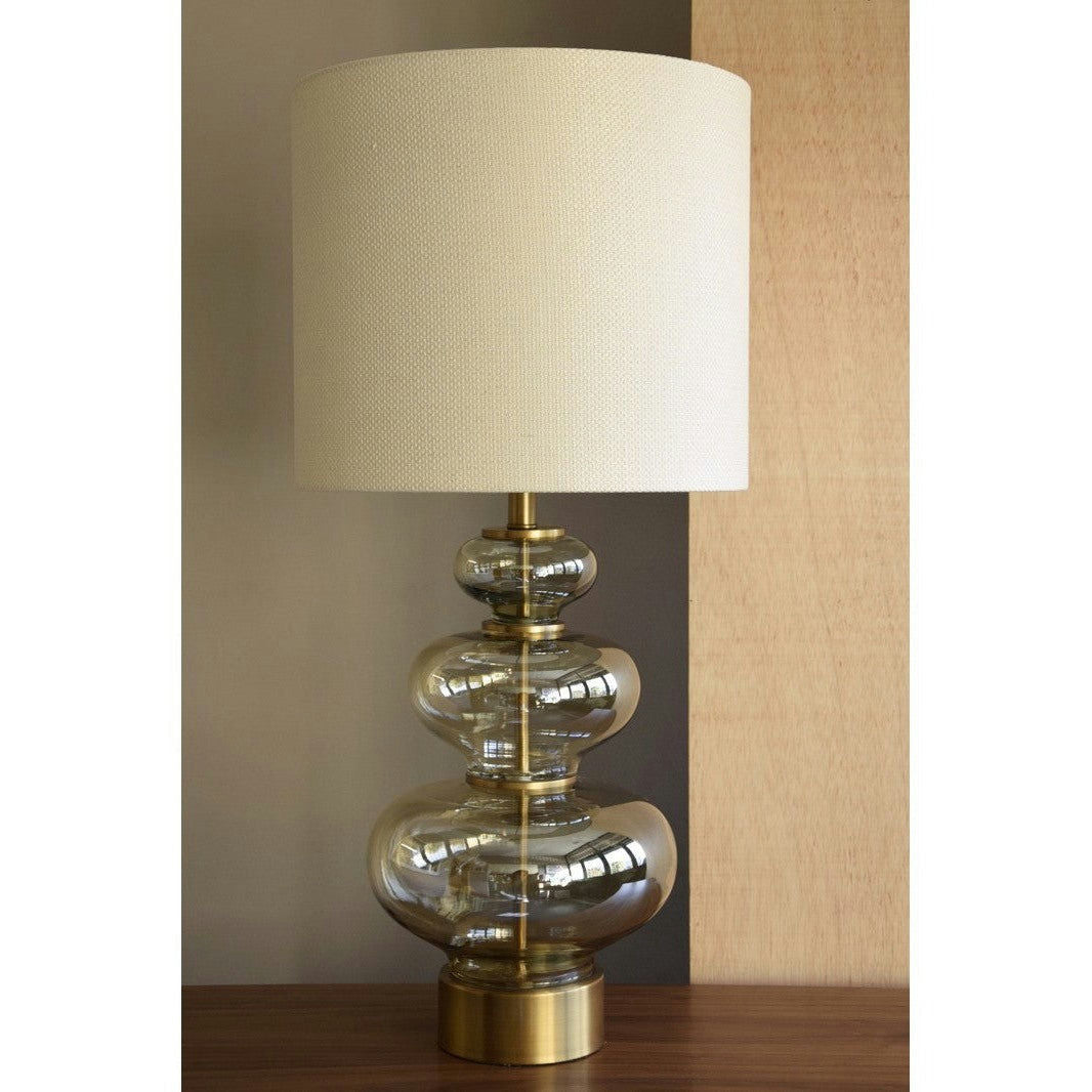 La Lámpara de Mesa de Vidrio Ámbar Calabaza cuenta con un diseño moderno con una base de vidrio transparente que presenta formas redondeadas apiladas. Su pantalla cilíndrica beige complementa la base envejecida en oro, lo que la convierte en un elegante complemento para cualquier superficie de madera.