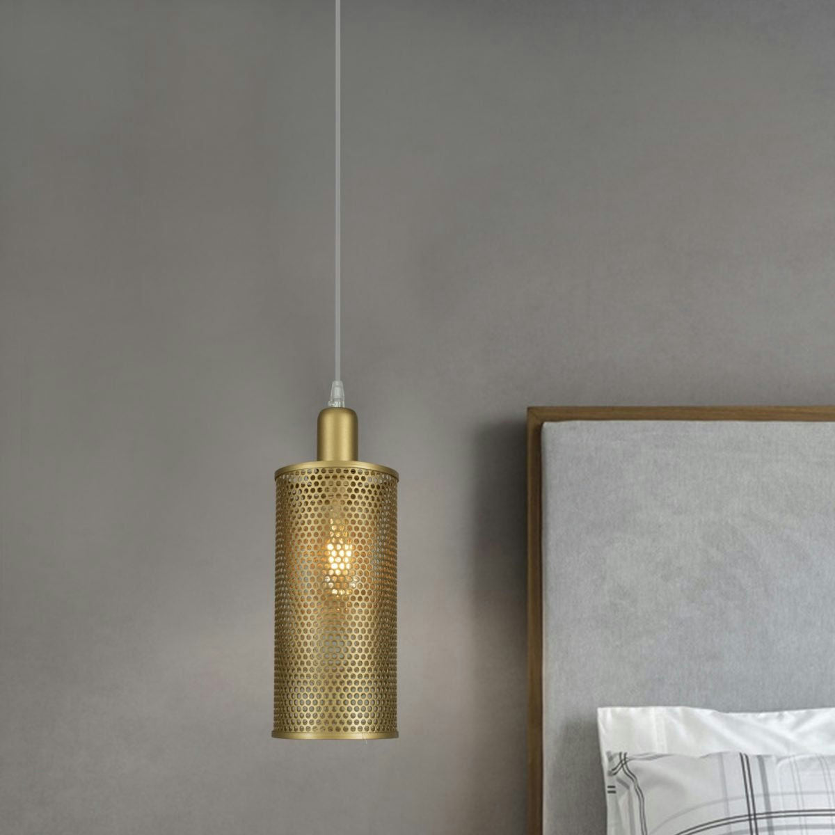 Una lámpara colgante cilíndrica dorada, la Lámpara de Techo Colgante Industrial Contemporáneo 3003-1C con un diseño perforado, cuelga junto a una cama tapizada en gris con ropa de cama blanca y gris.