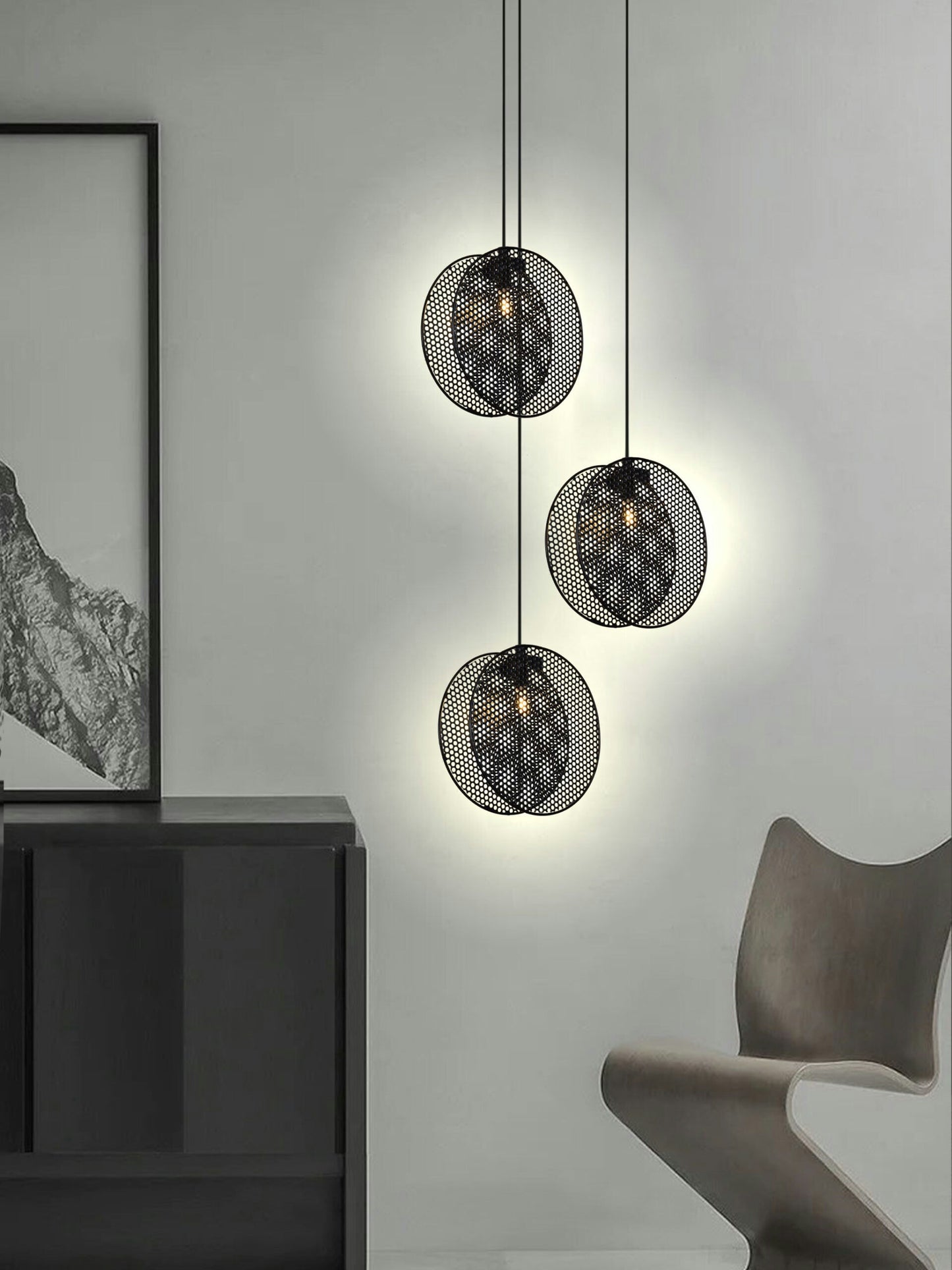Tres lámparas Lámpara de Techo Colgante Moderna 3224-3C-1 Negra con diseños perforados iluminan una pared gris claro sobre un gabinete oscuro y al lado de una silla moderna, agregando un estilo industrial chic a la habitación.