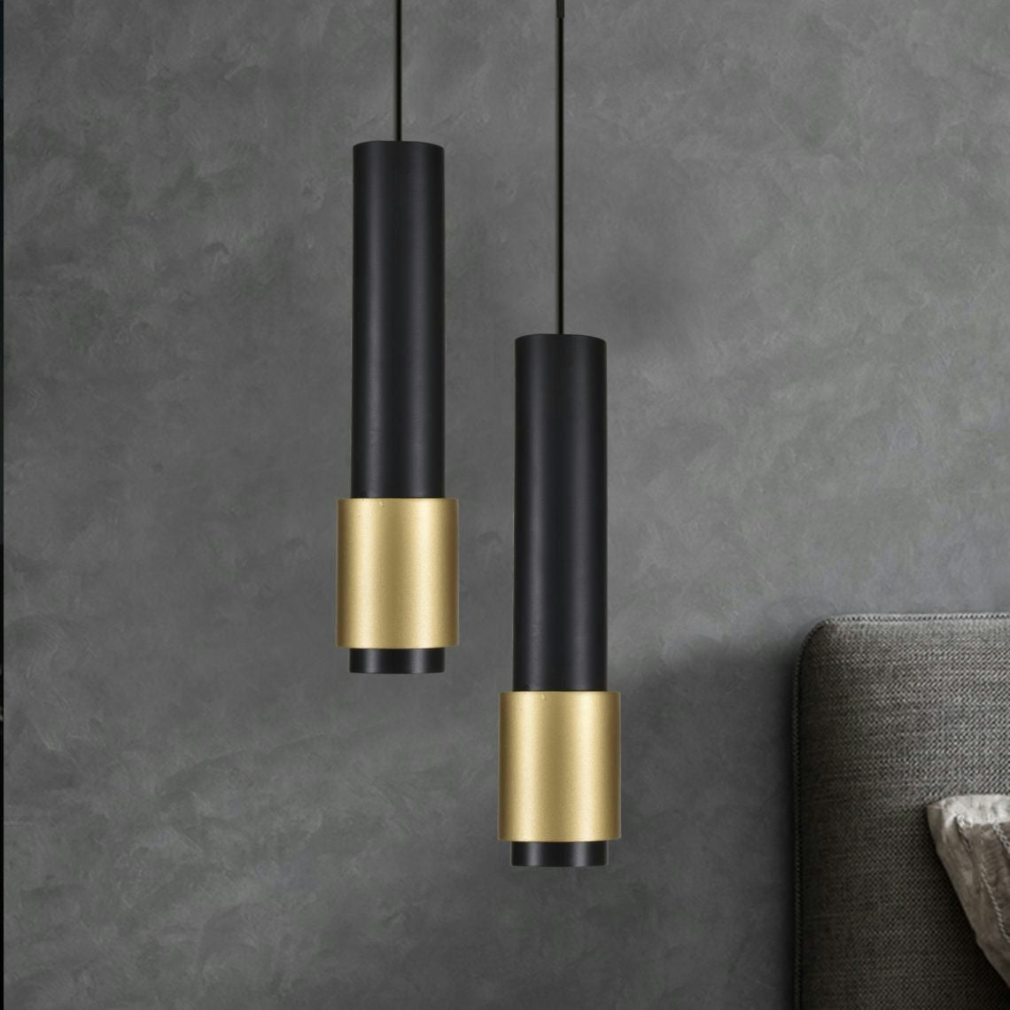 Dos elegantes y contemporáneas luces colgantes cilíndricas, la Lámpara de Techo Moderna Dorada y Negra 3044, cuelgan con estilo en acabados negros y dorados contra una pared gris texturizada sobre una parte de una cabecera acolchada.