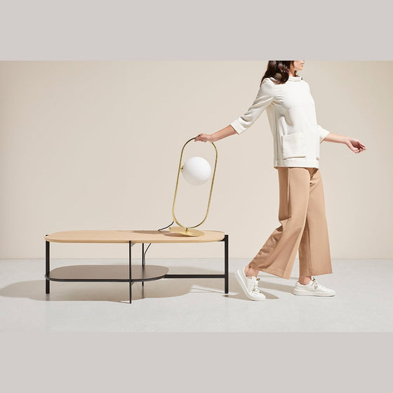 Una mujer con pantalones beige y una encimera blanca pasa junto a una mesa moderna con una Lámpara de mesa Abbacus sobre un fondo neutro, que refleja la elegancia contemporánea.