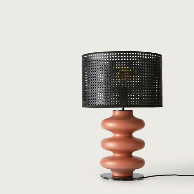 La lámpara de mesa Adon presenta una base ondulada de color terracota y una pantalla cilíndrica perforada en negro, que se alza elegantemente sobre un fondo blanco. Representa el diseño orgánico y ofrece iluminación moderna.