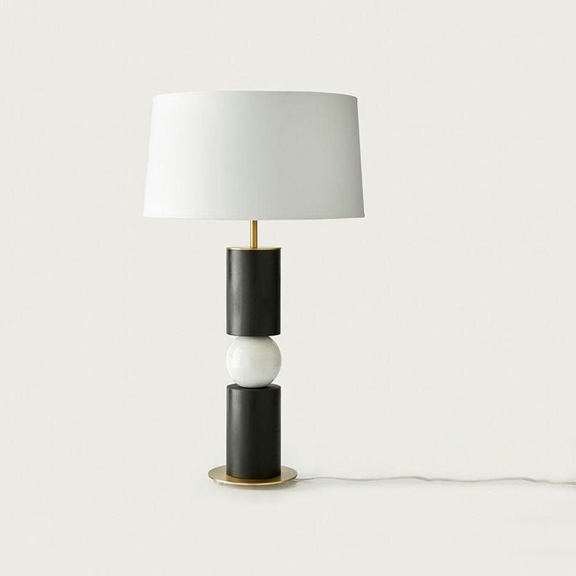 La lámpara de mesa Alda presenta una moderna pantalla cilíndrica blanca con un diseño contemporáneo, que incluye una base geométrica hecha de cilindros negros y una esfera blanca central, todo sobre una elegante base redonda dorada.