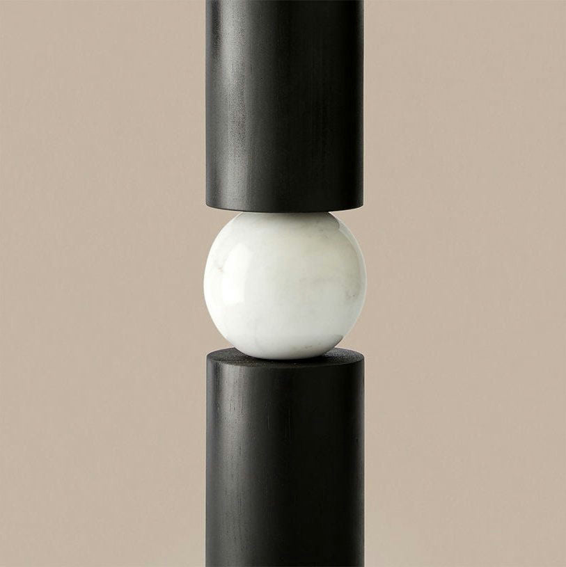La lámpara de mesa Alda presenta una esfera de mármol blanco elegantemente colocada entre dos columnas cilíndricas verticales negras, que irradia un estilo contemporáneo sobre un fondo beige, con sutiles toques de acabado dorado mate.