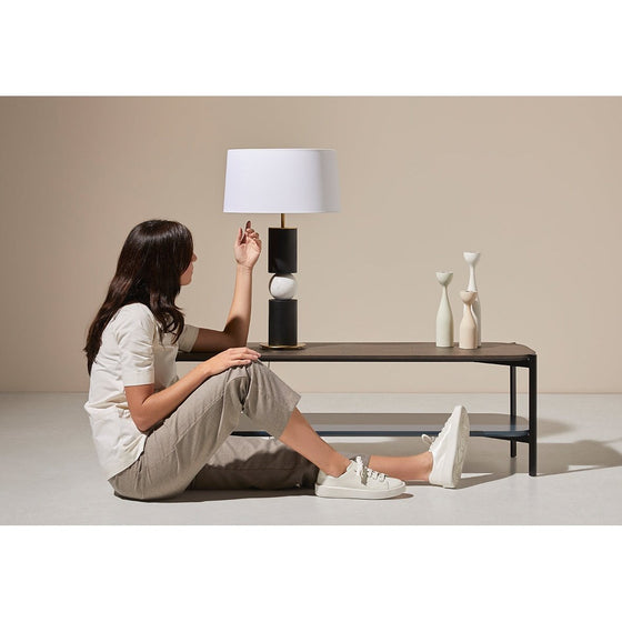 Mujer con ropa informal sentada en el suelo y tocando la Lámpara de mesa Alda, con jarrones decorativos cerca en un ambiente de tonos neutros y diseño contemporáneo.