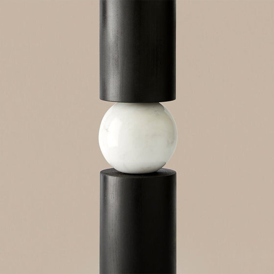 Una esfera de mármol blanco en equilibrio entre dos cilindros verticales negros sobre fondo beige, que evoca una Lámpara de mesa Alda contemporáneo.