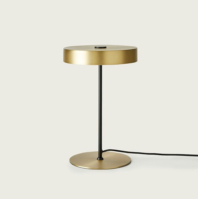 La lámpara de mesa Ambor presenta una pantalla cilíndrica dorada y una base conectadas por un fino tallo negro, que captura la iluminación minimalista sobre un fondo liso. Un elegante cable negro se extiende desde la base, personificando el diseño moderno.