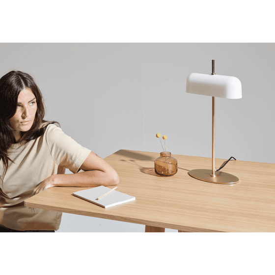 Una mujer sentada ante una mesa de madera con una lámpara de mesa Atil, una libreta y un accesorio de cristal, mirando pensativamente hacia un lado.