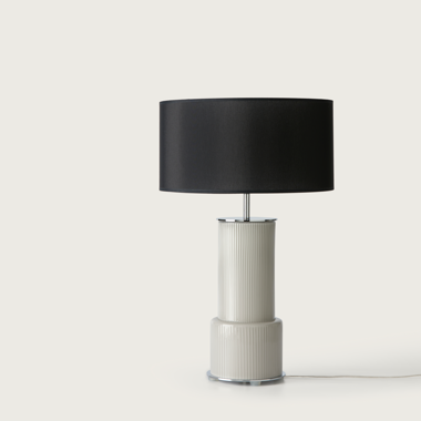 La lámpara de mesa Atina presenta una base de cerámica blanca texturizada y una pantalla cilíndrica de tela negra, ofreciendo un diseño clásico para crear un ambiente acogedor sobre un fondo claro.