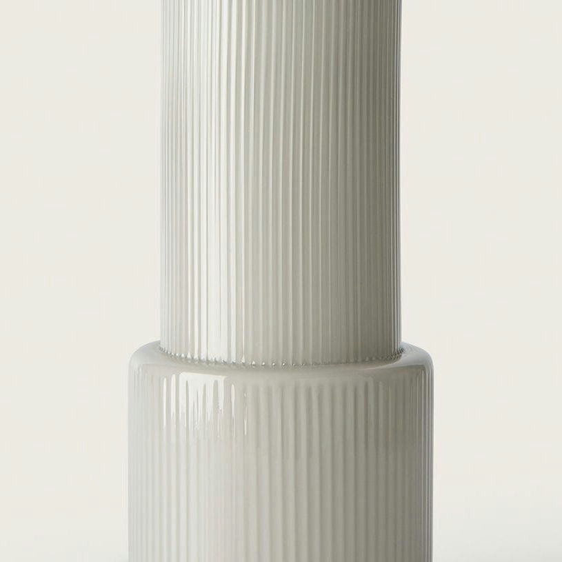 Primer plano de una lámpara de mesa Atina alta, de cerámica blanca, con textura acanalada sobre un fondo liso, añadiendo un toque de diseño clásico para crear un ambiente acogedor.