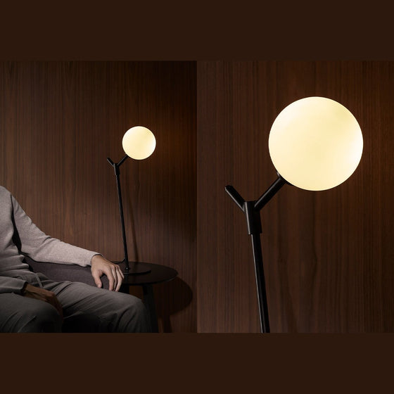 Una persona sentada junto a una elegante y moderna lámpara de mesa Atom con una gran luz esférica brillante, en una habitación con paredes de madera oscura.