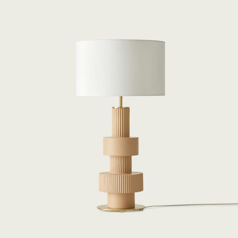 La lámpara de mesa Babel presenta una base de cerámica texturizada de color bronce y una elegante pantalla cilíndrica blanca, creando una impresionante pieza de arte decorativo que se destaca elegantemente sobre un fondo sencillo.